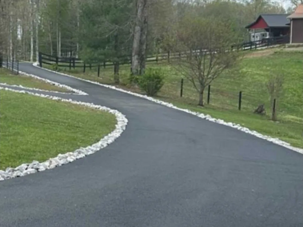 Curved asphalt driveway paving edged with white rock beside a fenced pasture