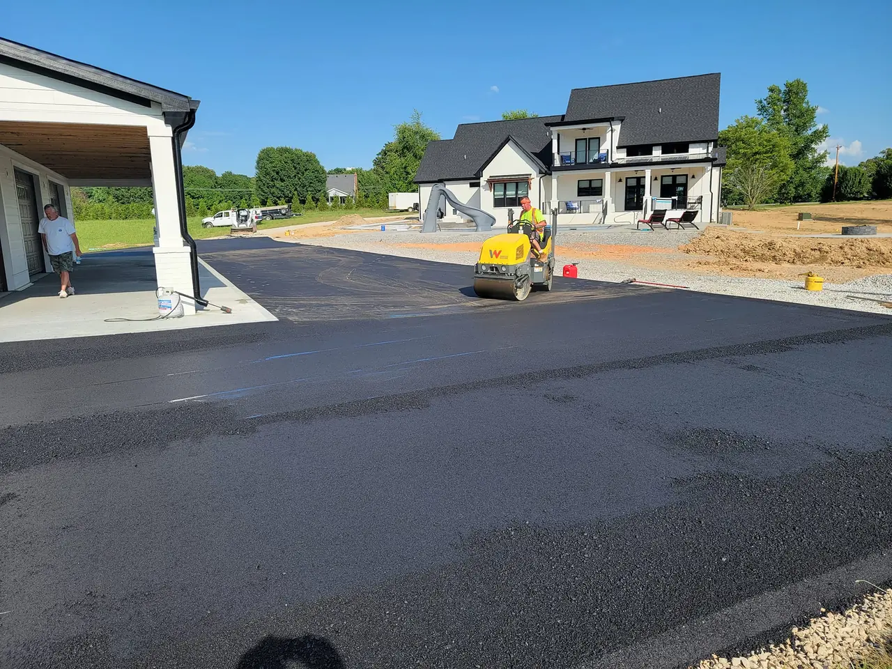 Asphalt driveway paving at a new white home with a roller compacting fresh hot mix