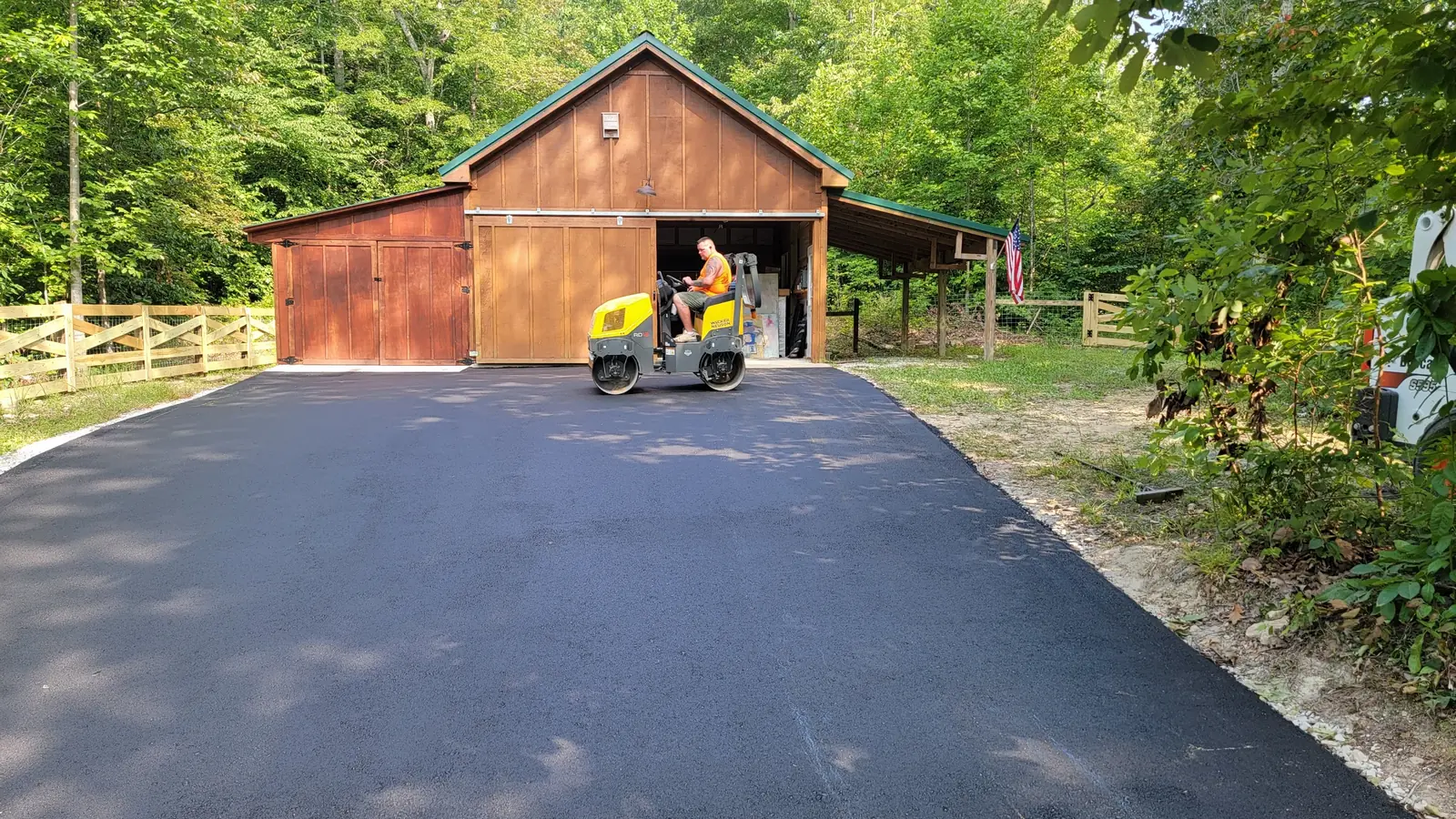 Asphalt paving contractor rolling a new asphalt pad in front of a wooden barn