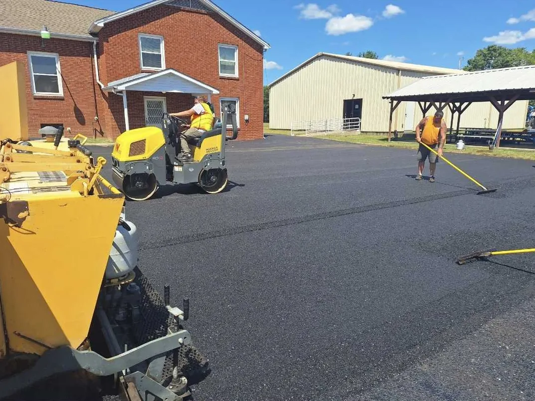 Parking lot paving crew raking fresh asphalt while a roller compacts the lot