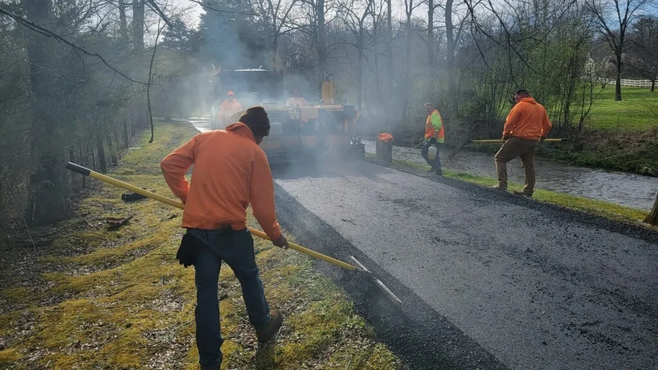 Asphalt paving crew raking hot mix behind a paver on a lane beside a creek
